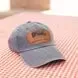 A light blue denim baseball cap with a brown rectangular leather patch on the front, sitting on a pink checkered surface.