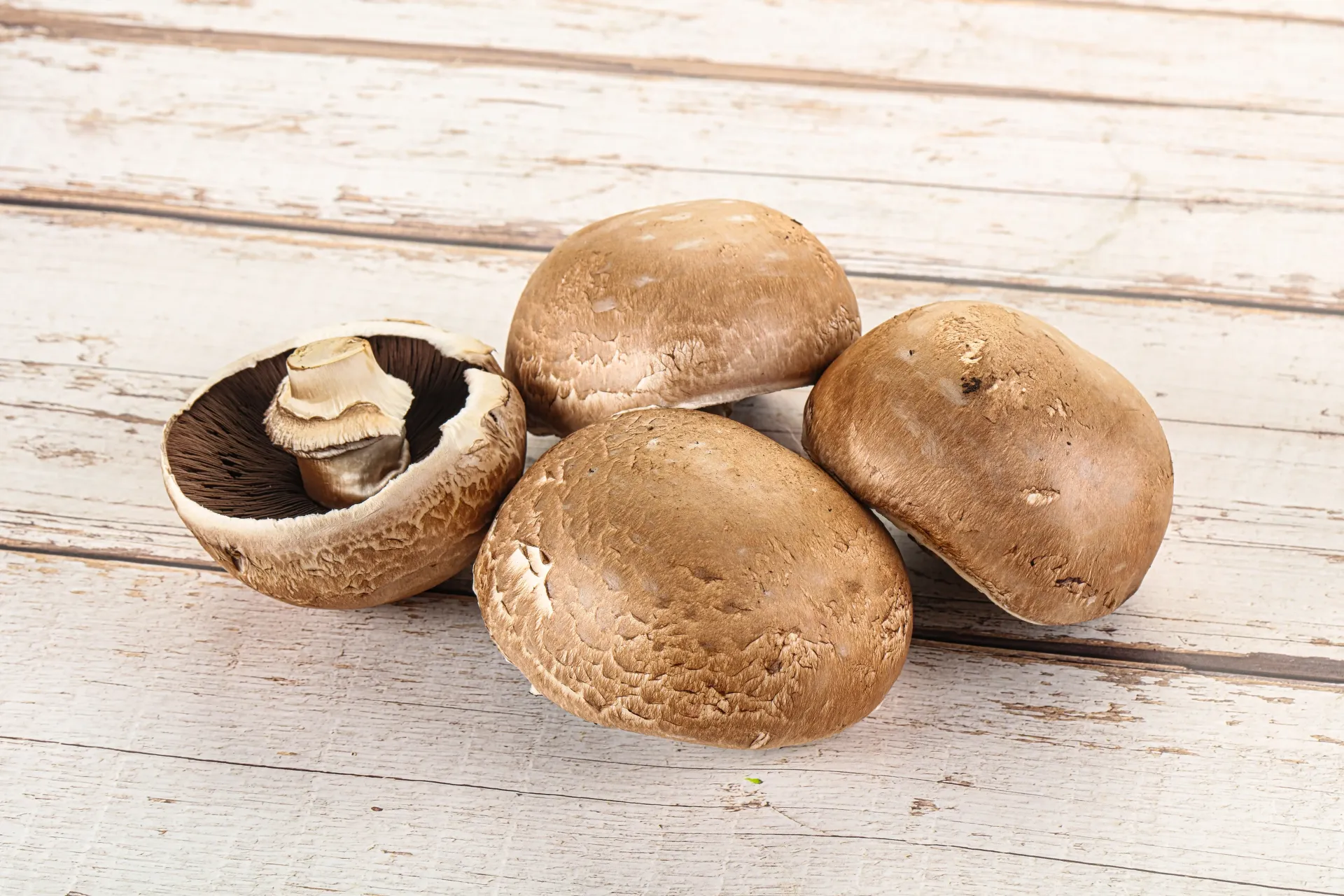 Four brown cremini mushrooms arranged on a light-colored wooden surface, with one mushroom turned upside down.