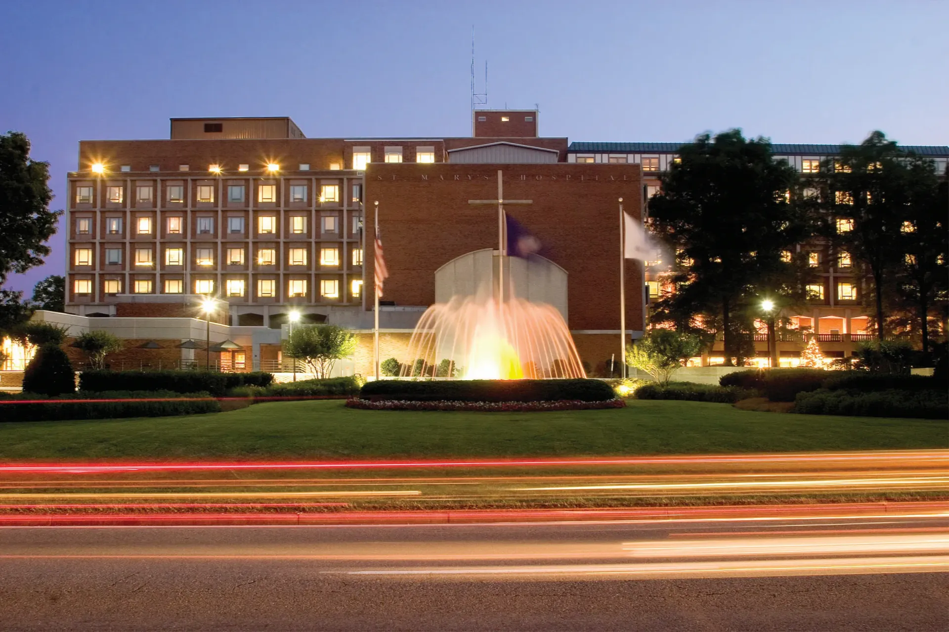 A multi-story brick hospital at dusk with a large illuminated fountain and flags in the foreground and light trails below.