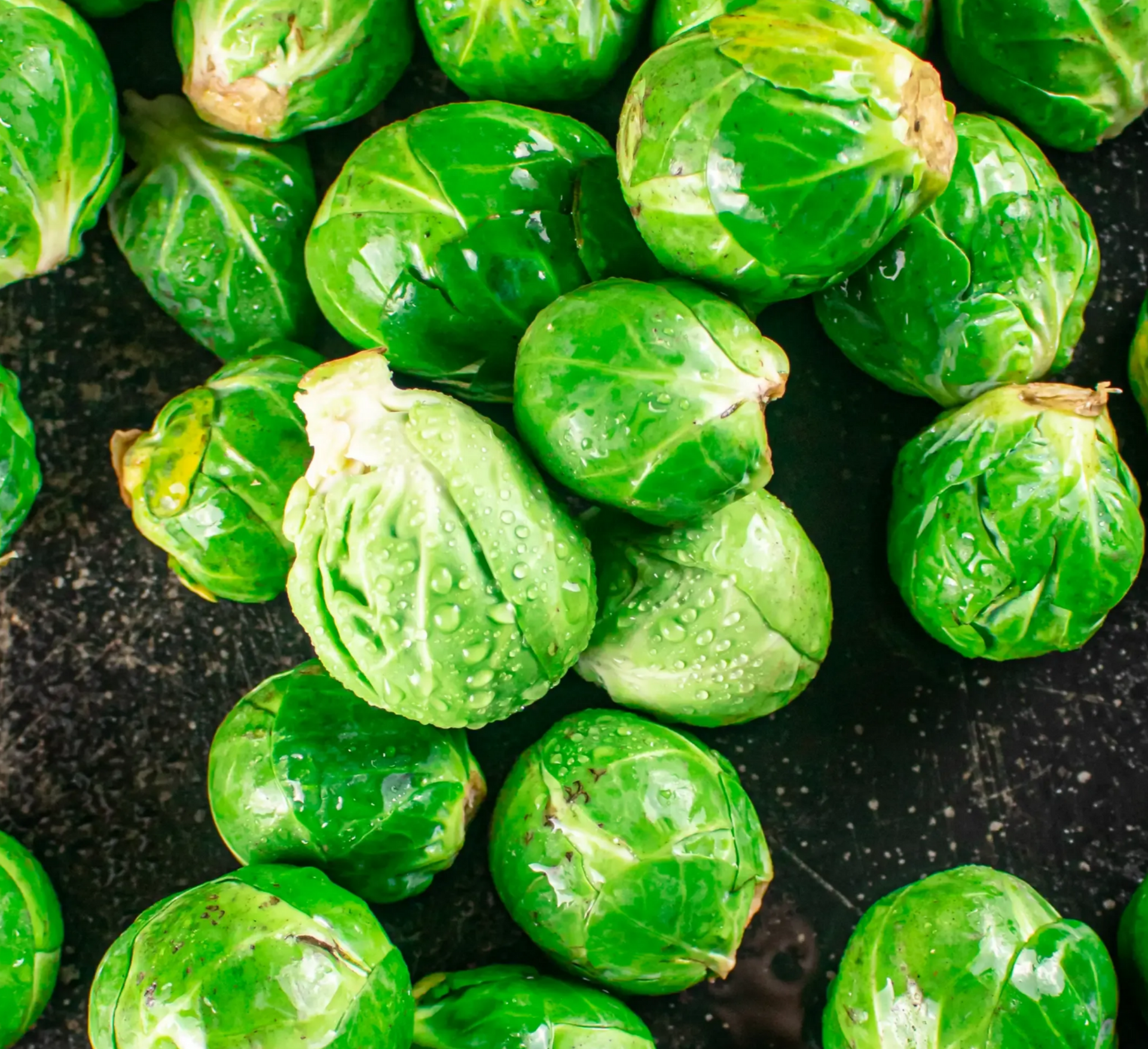 A pile of fresh, vibrant green Brussels sprouts with water droplets scattered on a dark, textured background.
