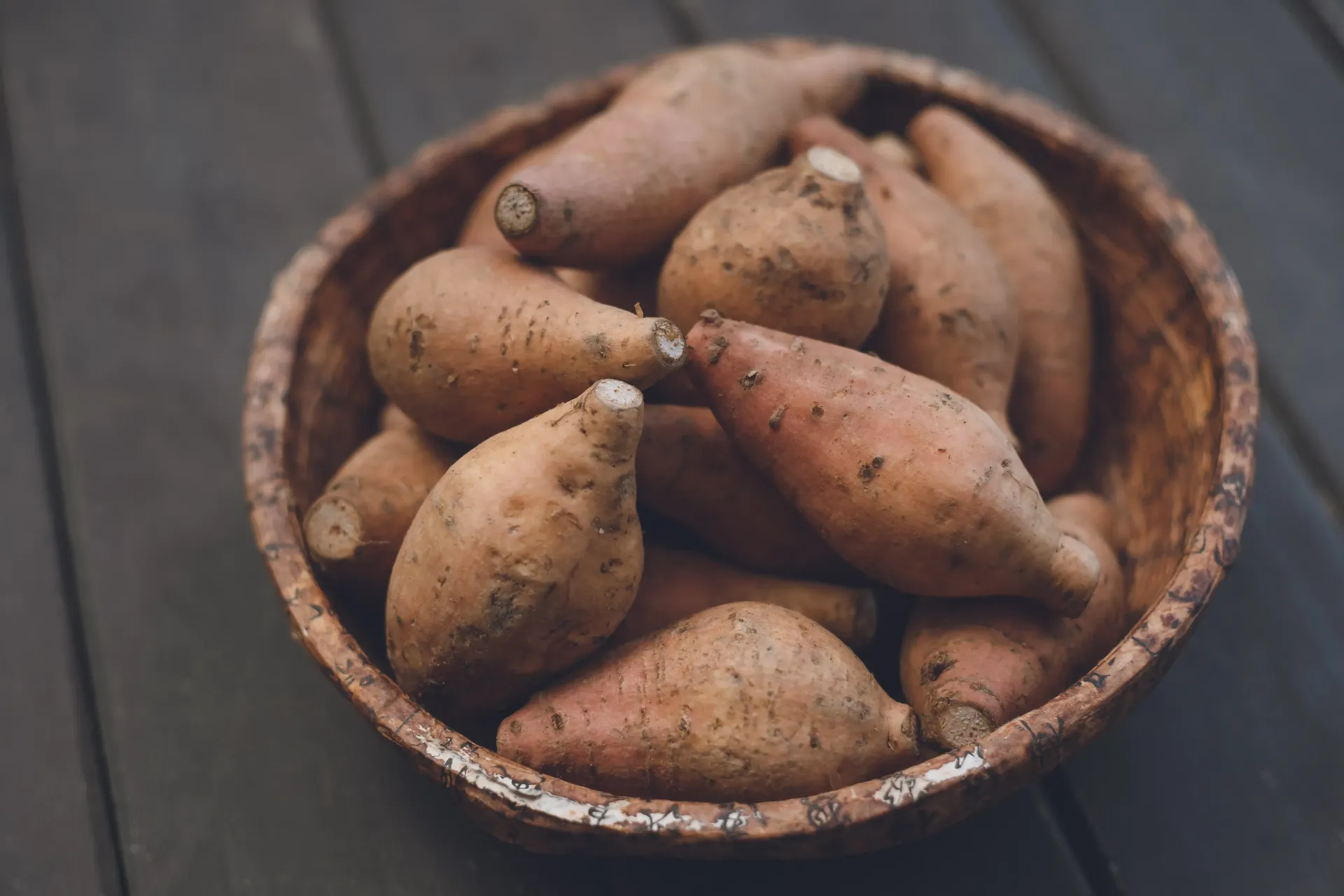 A rustic wooden bowl filled with several raw, unpeeled sweet potatoes on a dark wooden surface.