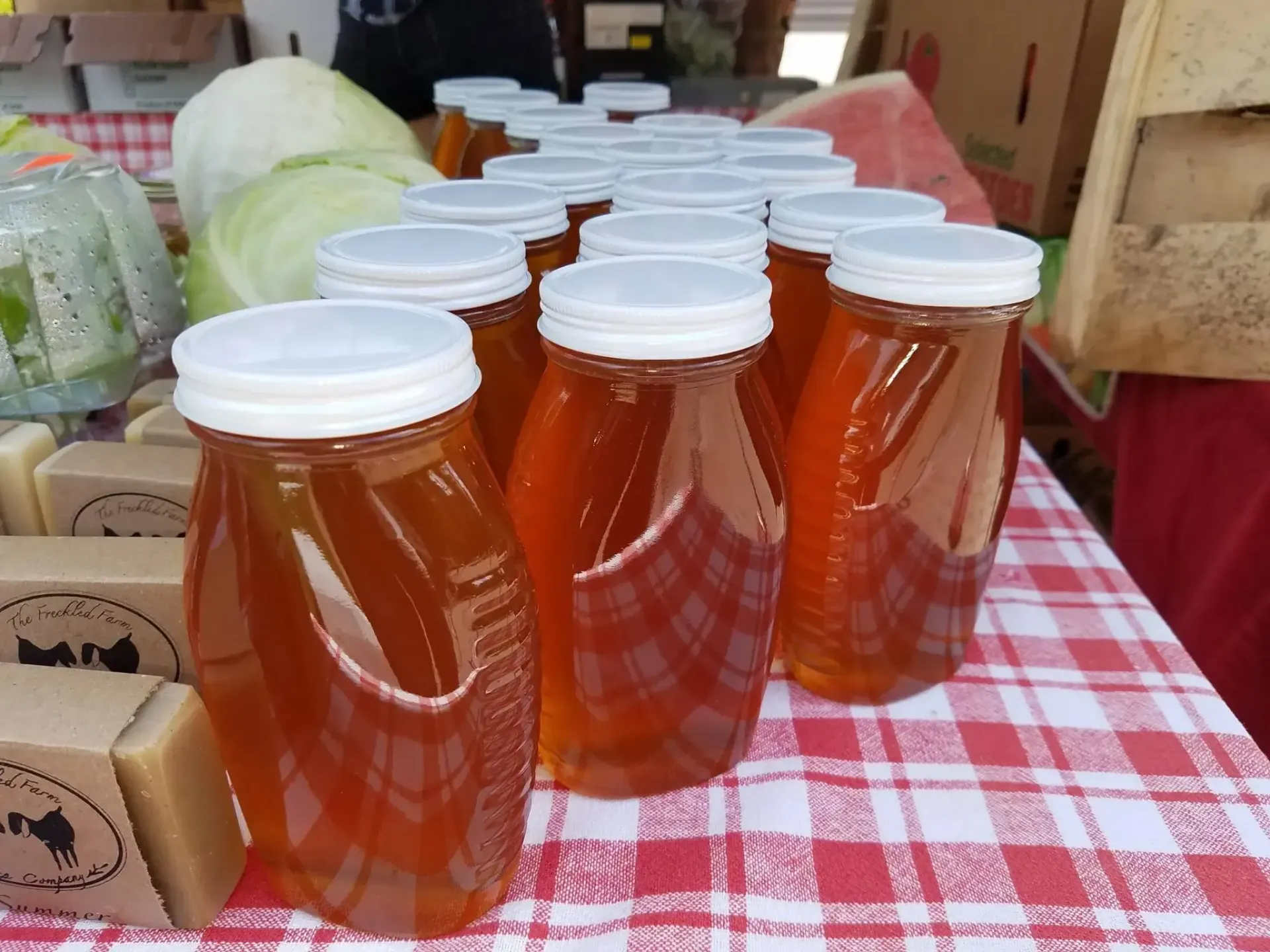 Several glass jars of golden honey with white lids sit on a red and white checkered tablecloth next to bars of soap.