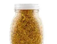 A plastic, hive-shaped squeeze bottle filled with golden-brown bee pollen granules, topped with a white screw-on cap.