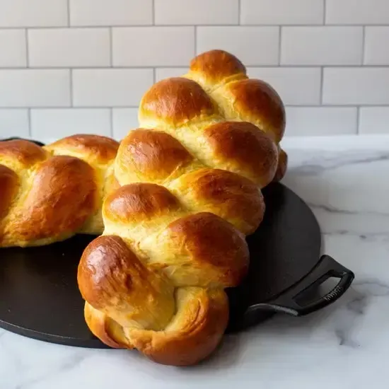 Two braided loaves of golden-brown challah bread sit on a round black tray against a white subway tile backsplash.