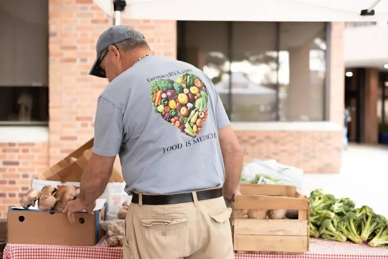 A person in a light blue shirt with a produce-filled heart design sorts fresh vegetables at an outdoor market stall.