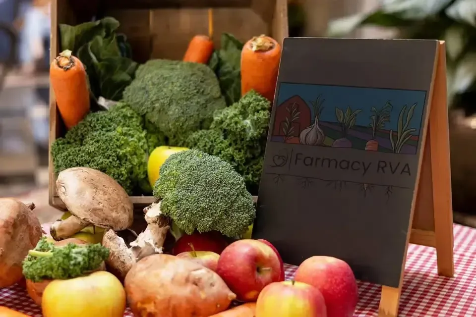 A crate filled with fresh broccoli, carrots, and mushrooms next to a Farmacy RVA sign on a red and white checkered cloth.