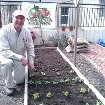 A smiling person kneels in front of a raised garden bed with small leafy plants, in front of a Virginia Grown banner.