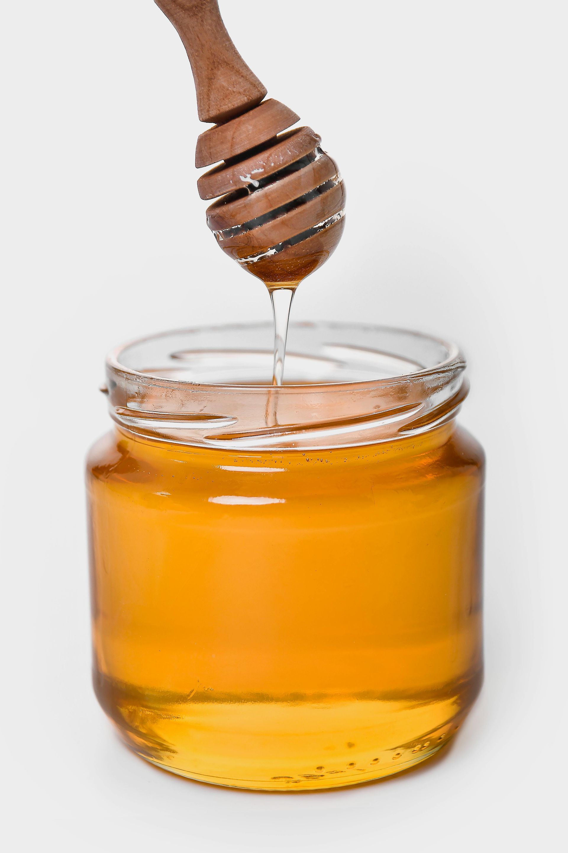 A honey dipper dripping golden honey into a clear glass jar against a white background.