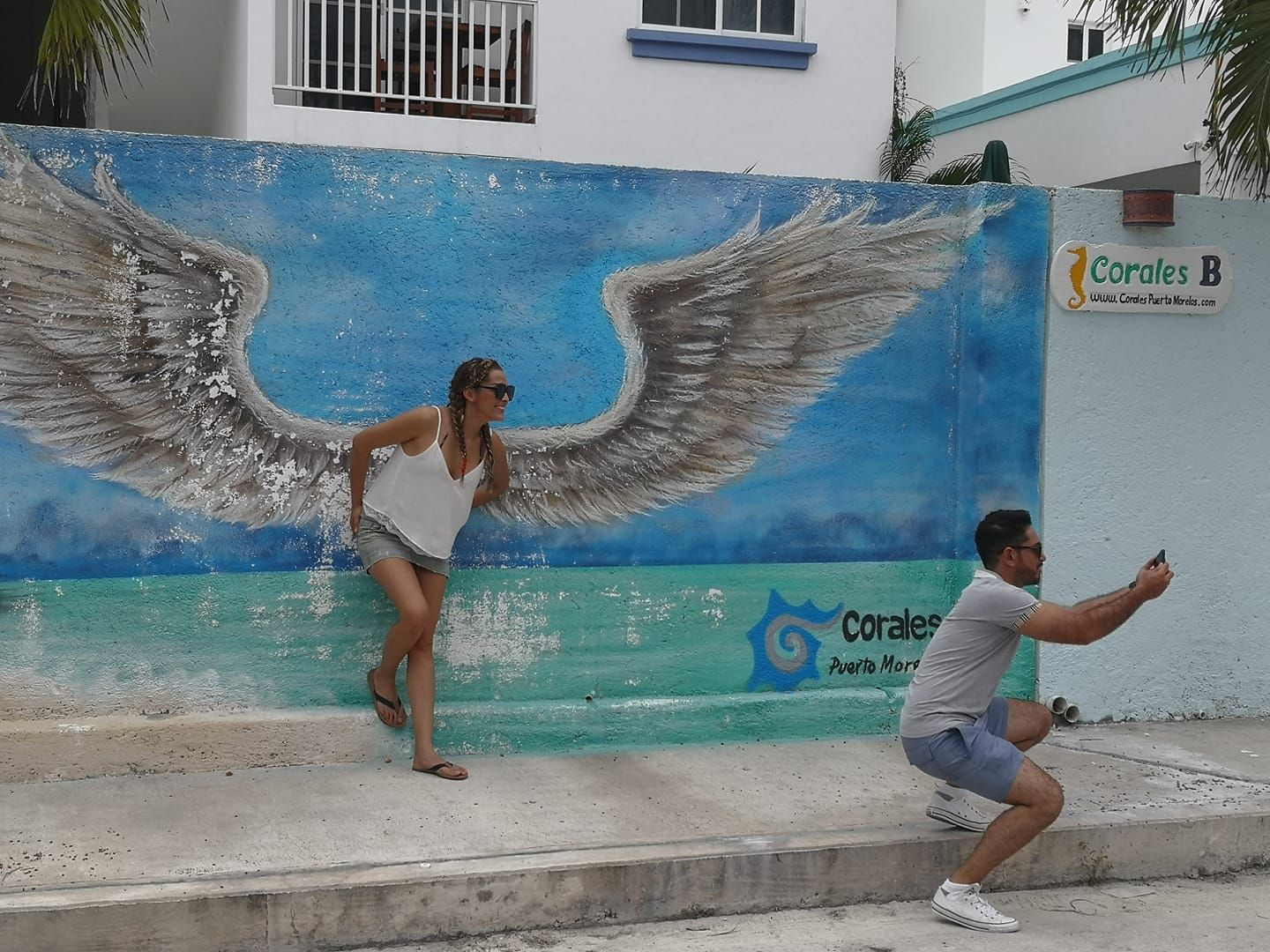 Mujer posando junto a un mural azul con alas de ángel mientras un hombre le toma una foto. Cielo azul claro.