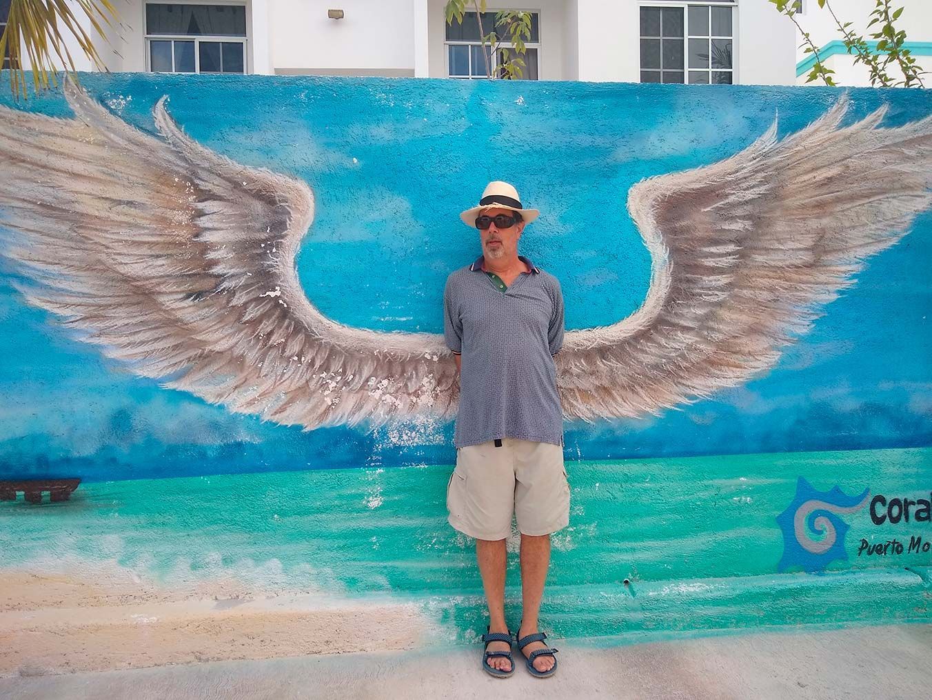 Man stands in front of a mural of angel wings painted on a blue wall, wearing a hat, shorts, and sandals.