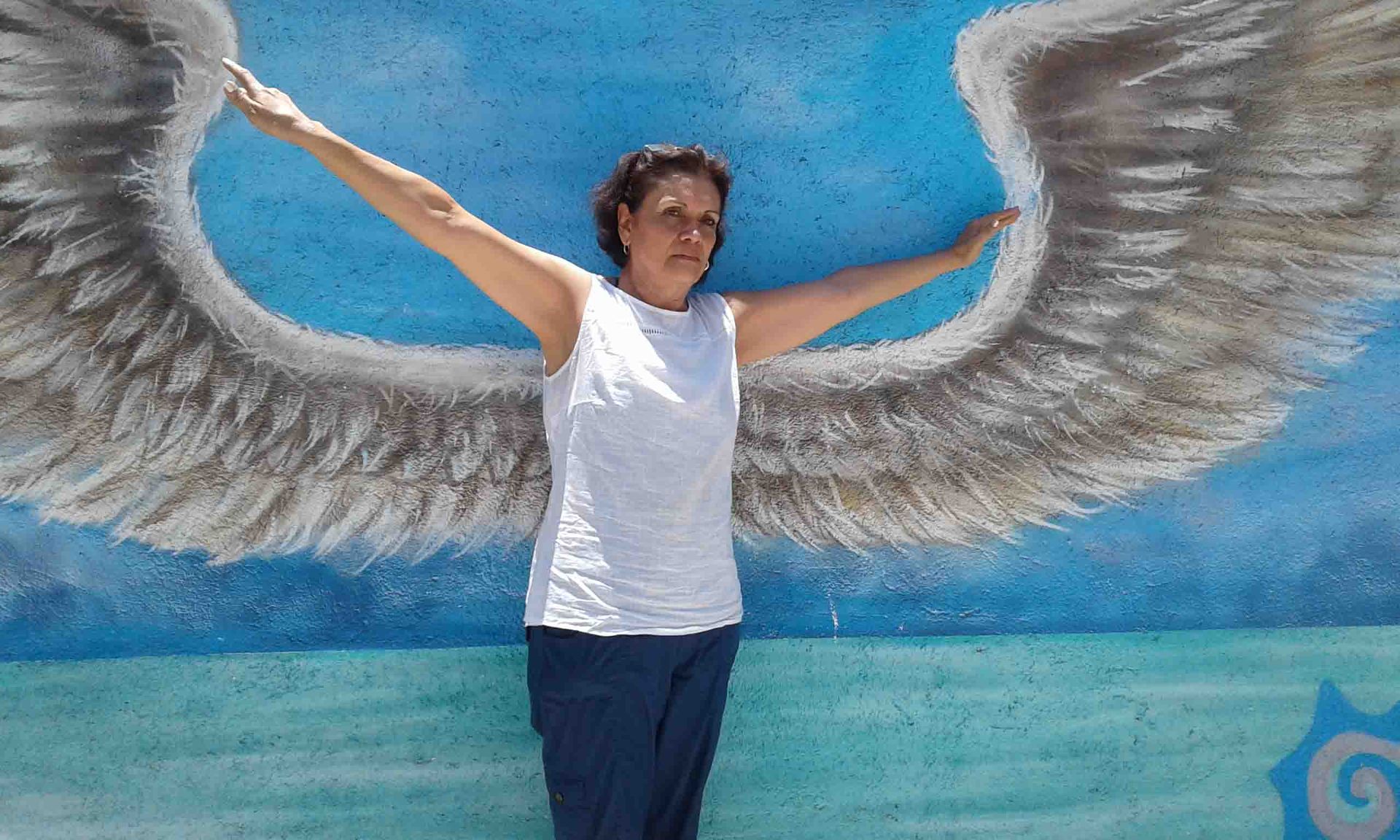 Woman posing with outstretched arms in front of a mural of angel wings painted on a wall.