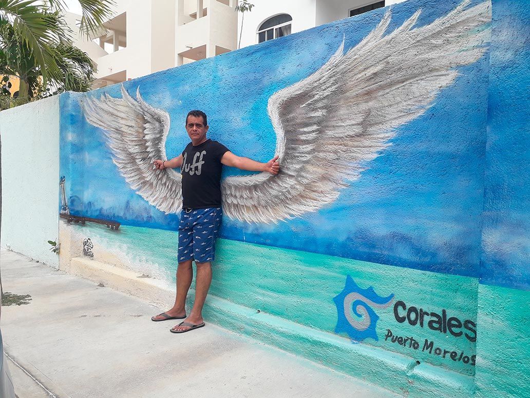 Un hombre posa con los brazos extendidos frente a un mural de alas de ángel en una pared azul en Puerto Morelos.
