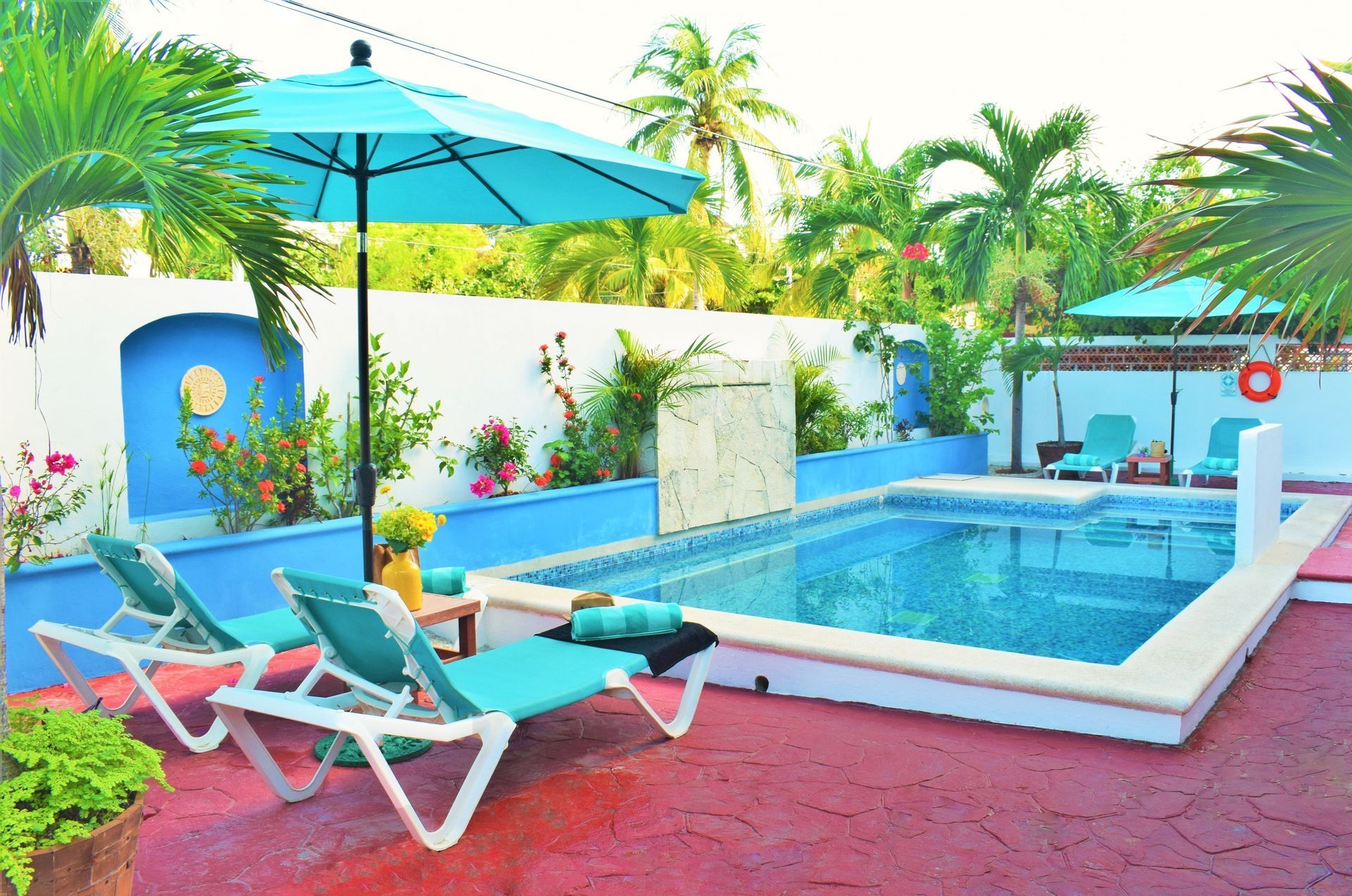 Escena junto a la piscina con sombrilla color turquesa, tumbonas y pared con flores.