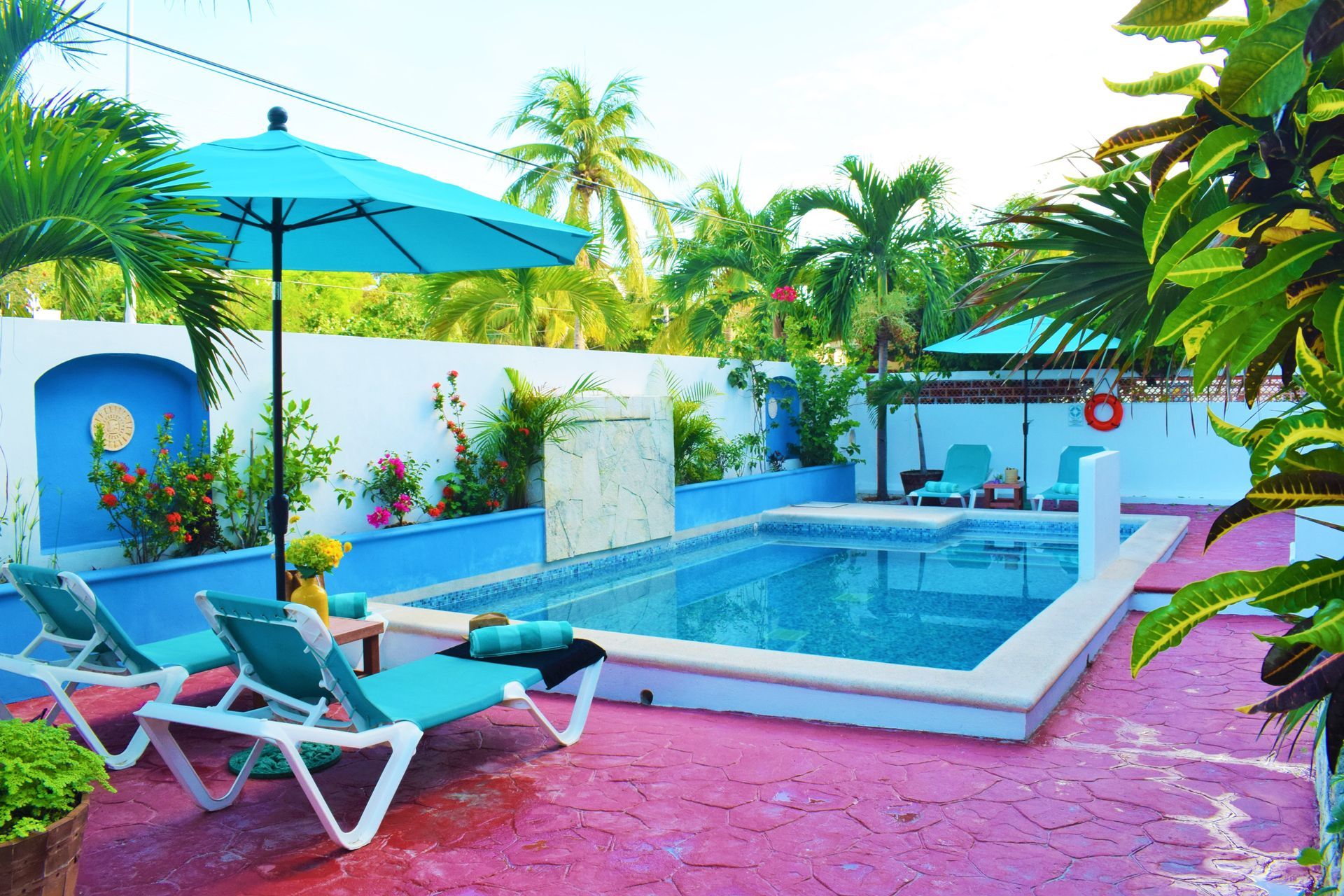 Escena junto a la piscina: sombrilla azul, tumbonas, piscina, palmeras, paredes blancas y azules, suelo rosa.