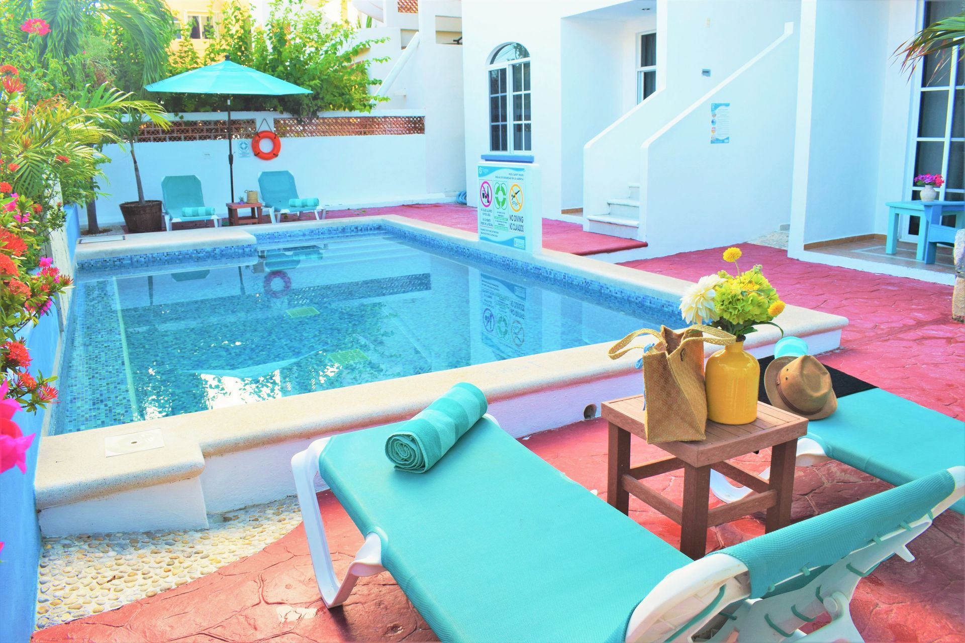 Escena junto a la piscina: tumbonas turquesas, una mesita con bebidas, piscina, edificios blancos y plantas.