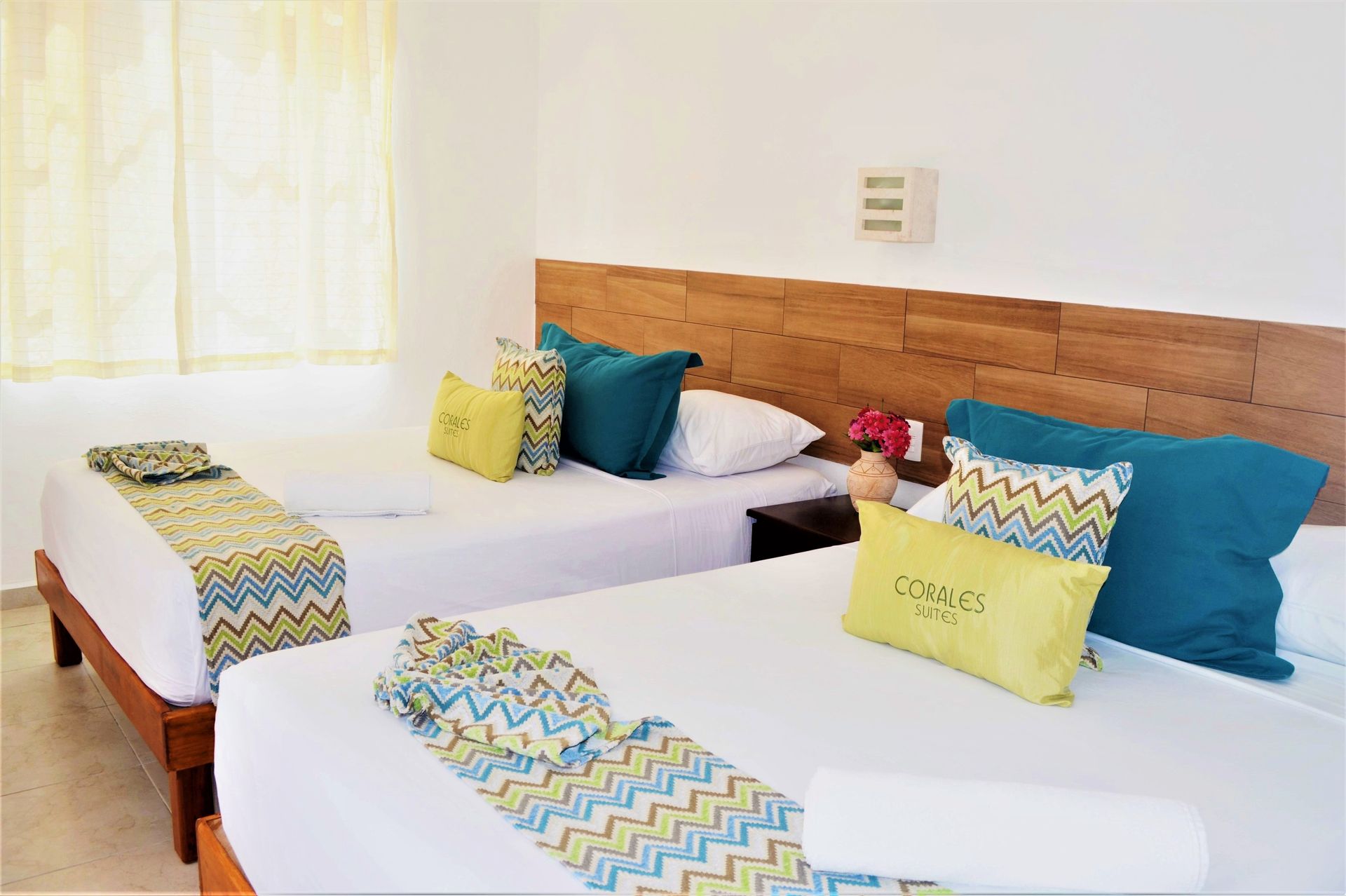 Two beds in a bright room with teal and yellow pillows and accents, wooden headboard.