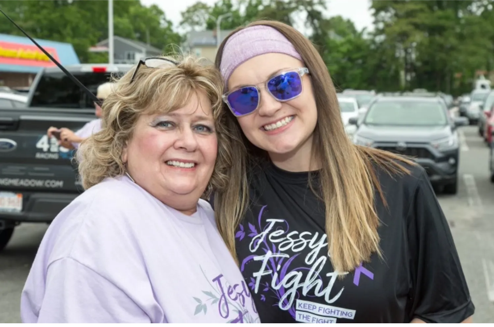 Two women are posing for a picture in a parking lot.