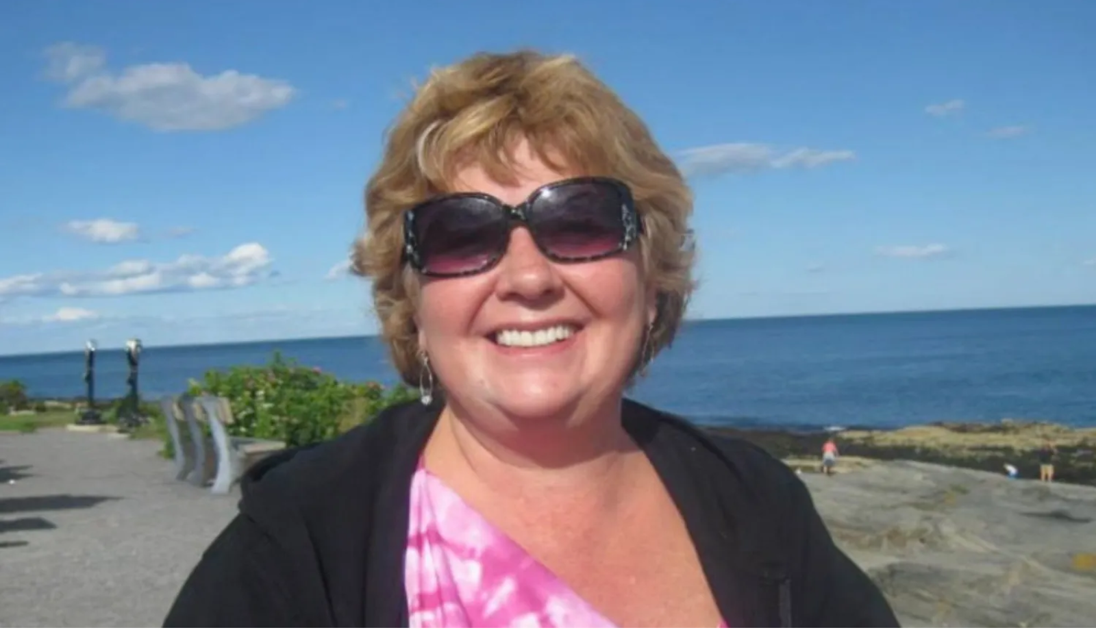 A woman wearing sunglasses is smiling in front of the ocean