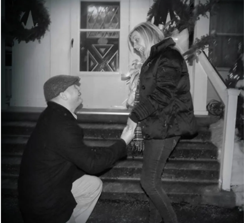 A man is kneeling down to propose to a woman on the steps of a house.