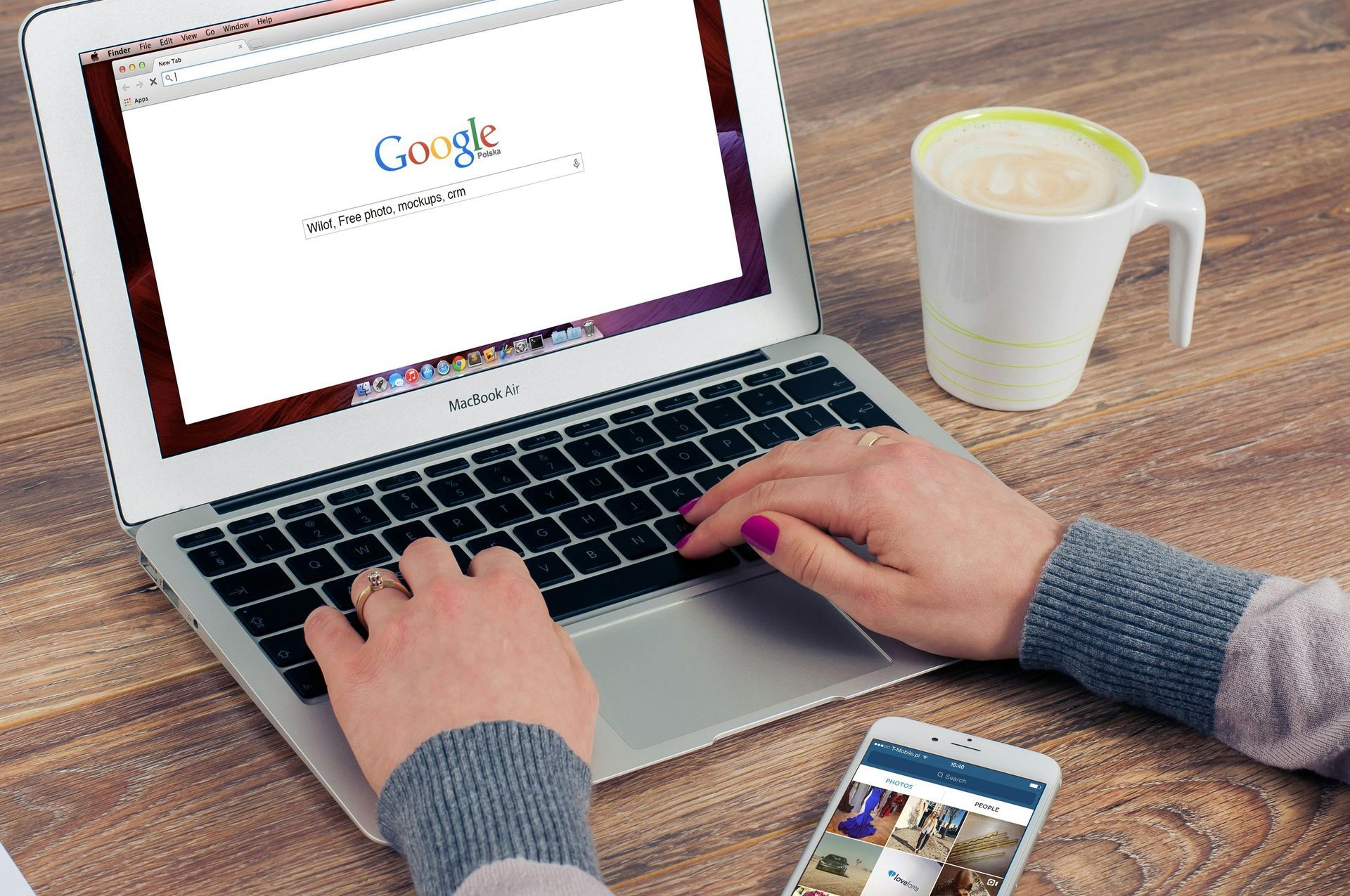 Hands typing on a laptop at a wooden desk with a coffee mug, smartphone, and Google open on screen