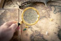 Hand holding a magnifying glass over an old map with a compass and parchment background
