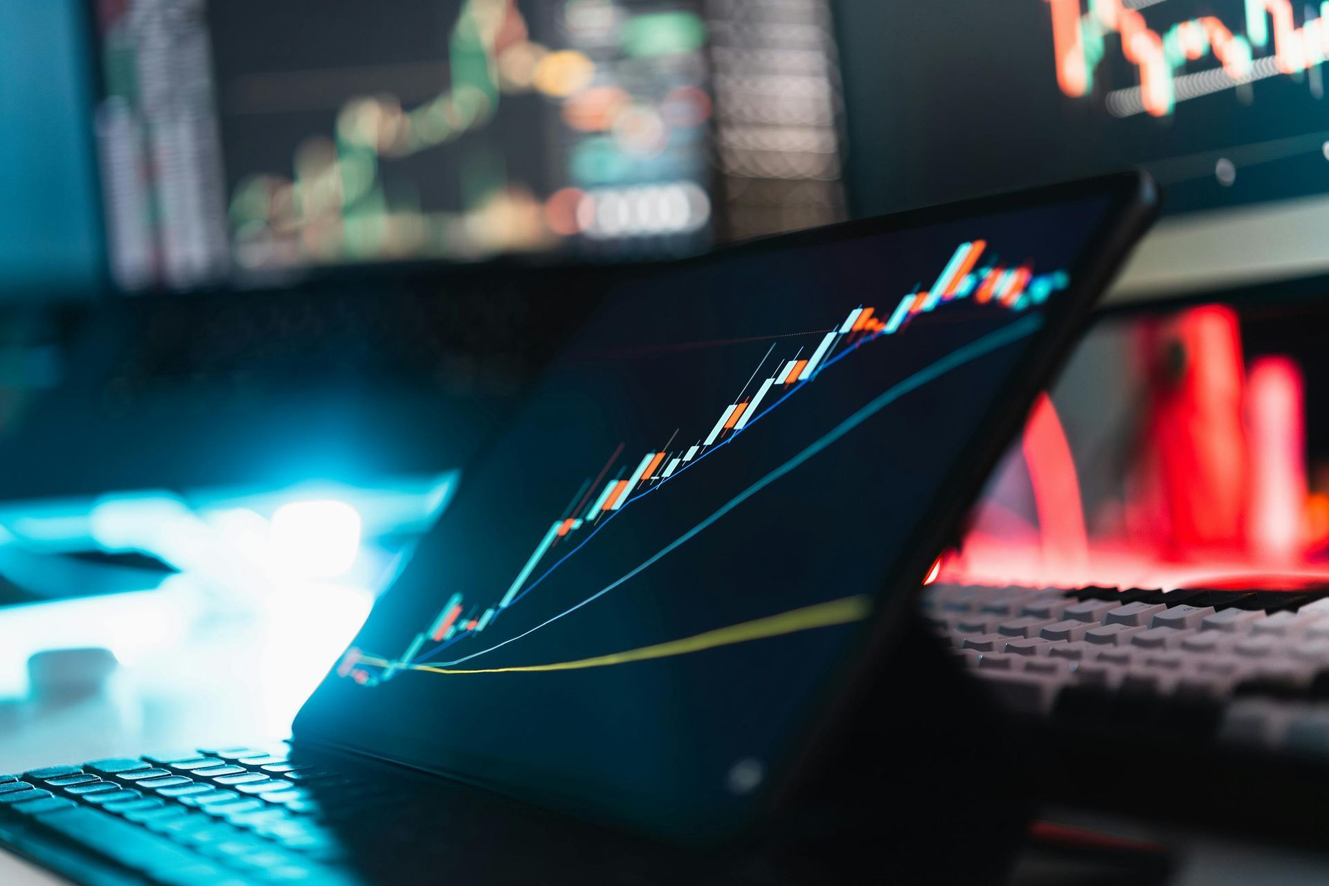 Laptop with glowing stock charts in a dim trading workspace, lit by blue and red screens