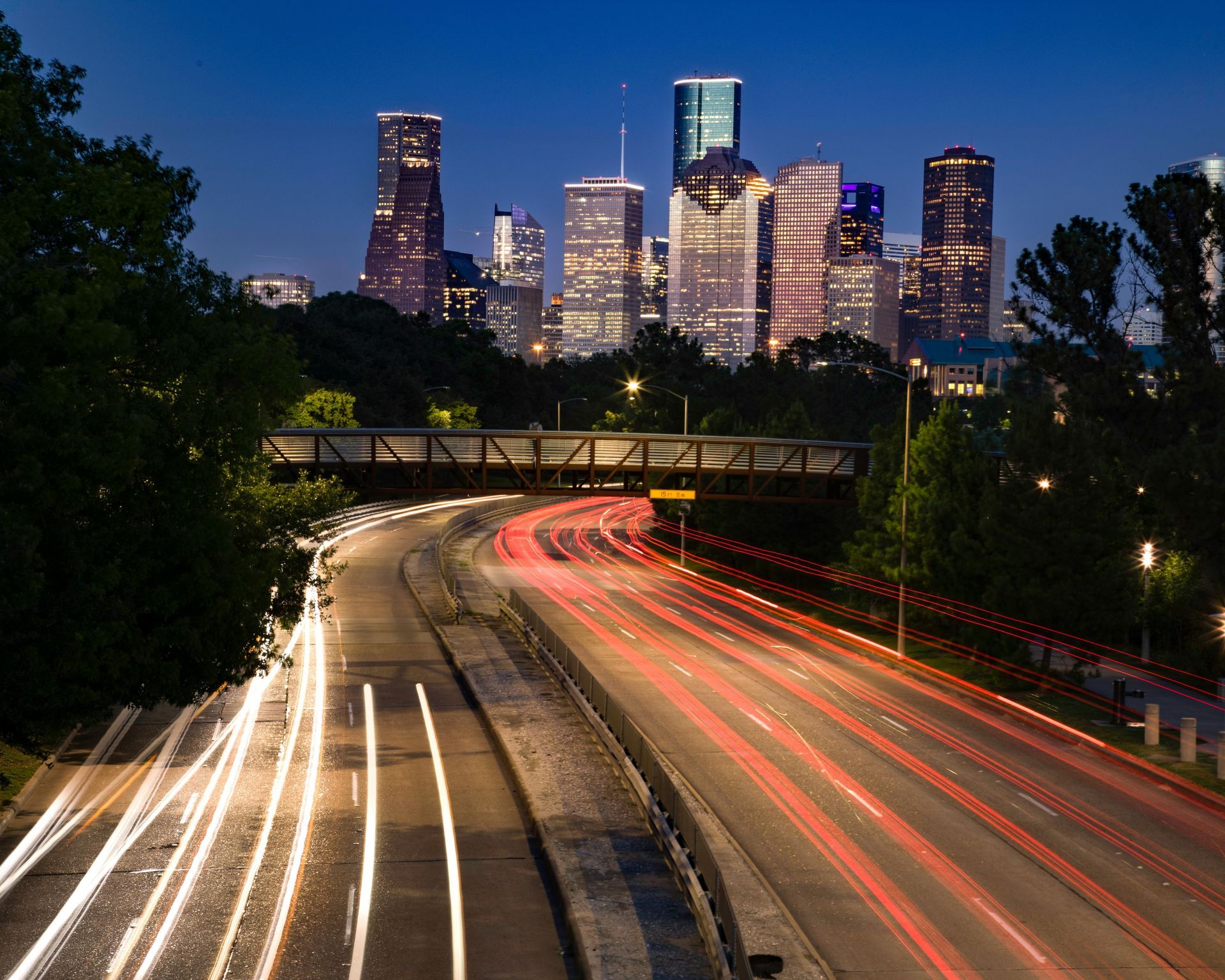 Nighttime city skyline beyond a highway, with red and white car light trails curving through trees