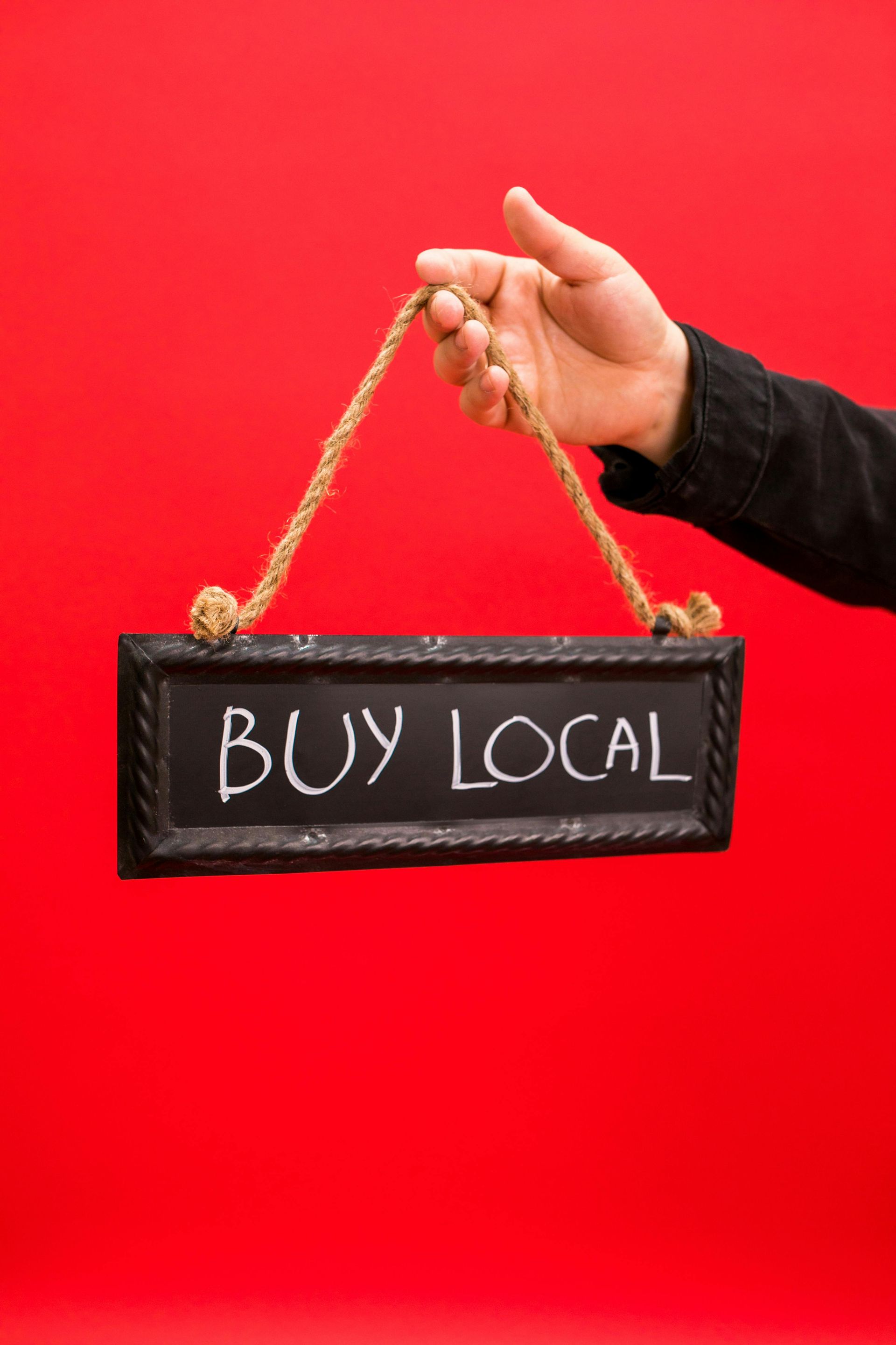 Hand holding a small chalkboard sign reading “BUY LOCAL” against a red background