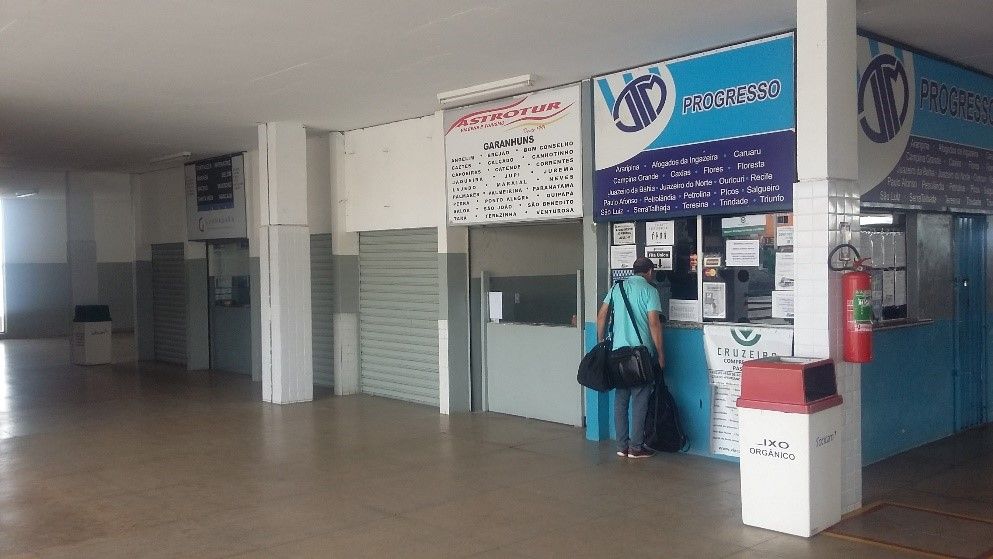 Táxis brancos enfileirados sob a cobertura de um prédio; plantas verdes e um céu azul se destacam.