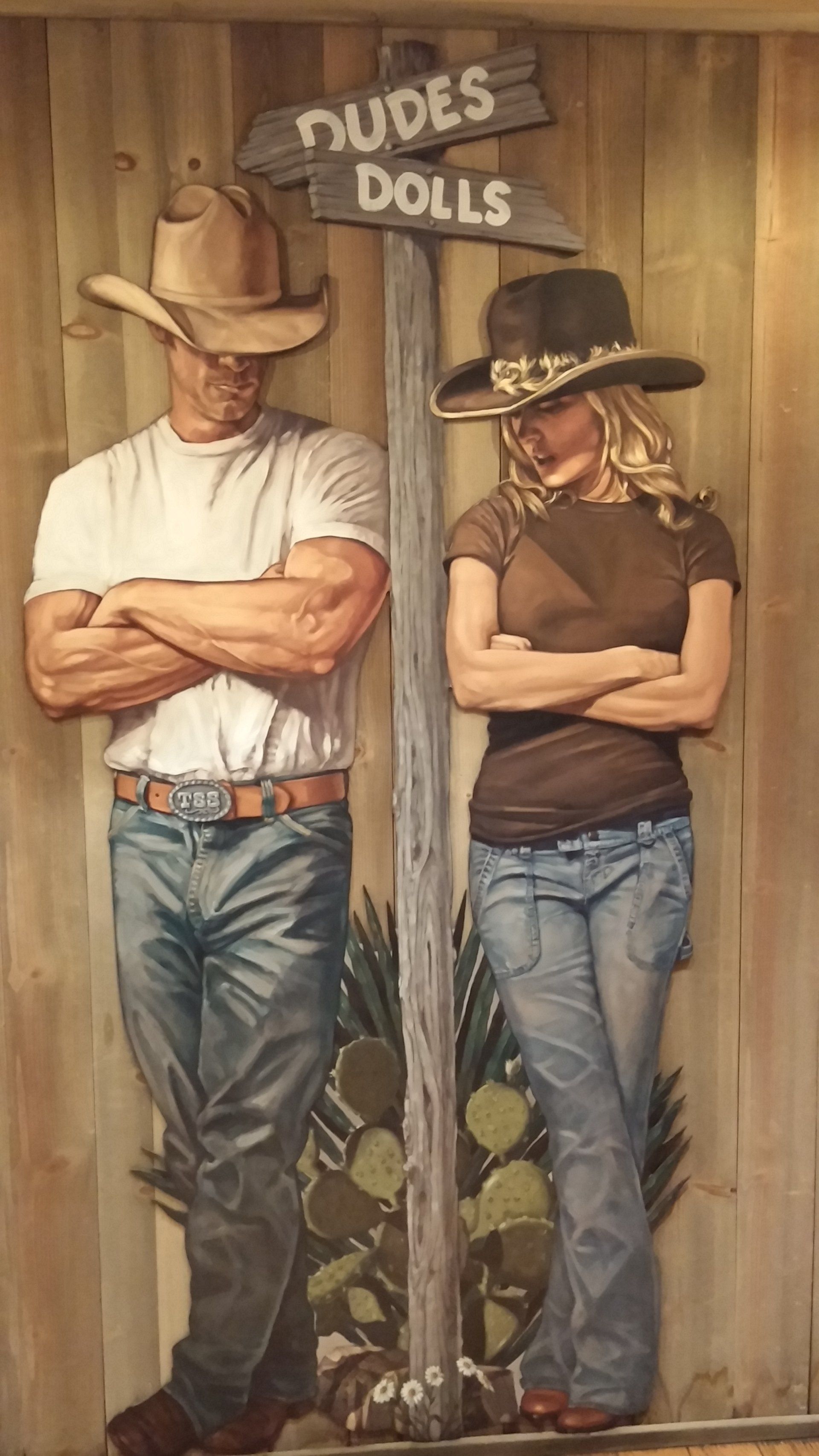 Cowboy and woman posing with arms crossed under a “Dudes/Dolls” sign. Wooden background.