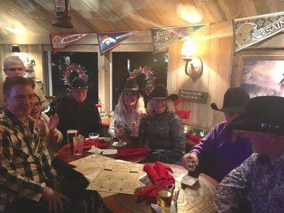 Group of people at a table in a rustic setting; some wearing cowboy hats and toasting with drinks.