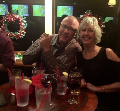 Man and woman smiling at a table with drinks; festive decorations in the background.