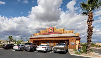 Restaurant with wooden facade and horse logo, cars parked in front. Palm tree and cloudy sky in background.