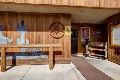 Entrance to a rustic Western-themed building with wooden facade, featuring a bench, wagon wheel, and cactus cutouts.