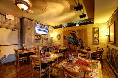 Interior of a rustic restaurant with wooden walls, tables, and chairs, featuring a rodeo-themed mural and a TV screen.