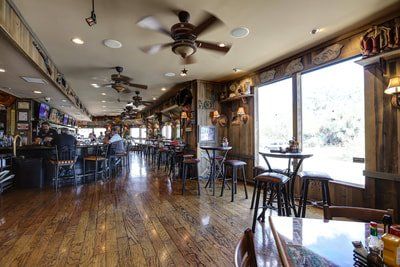 Interior of a bar with wooden floors, tables, bar stools, and patrons, featuring natural light and decor.