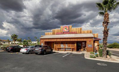 Restaurant with wooden facade and parking lot under a cloudy sky. Palm tree on the right.