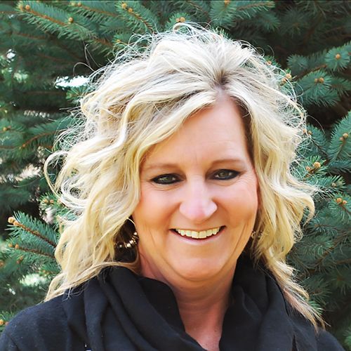 A woman with blonde hair is smiling in front of a pine tree.