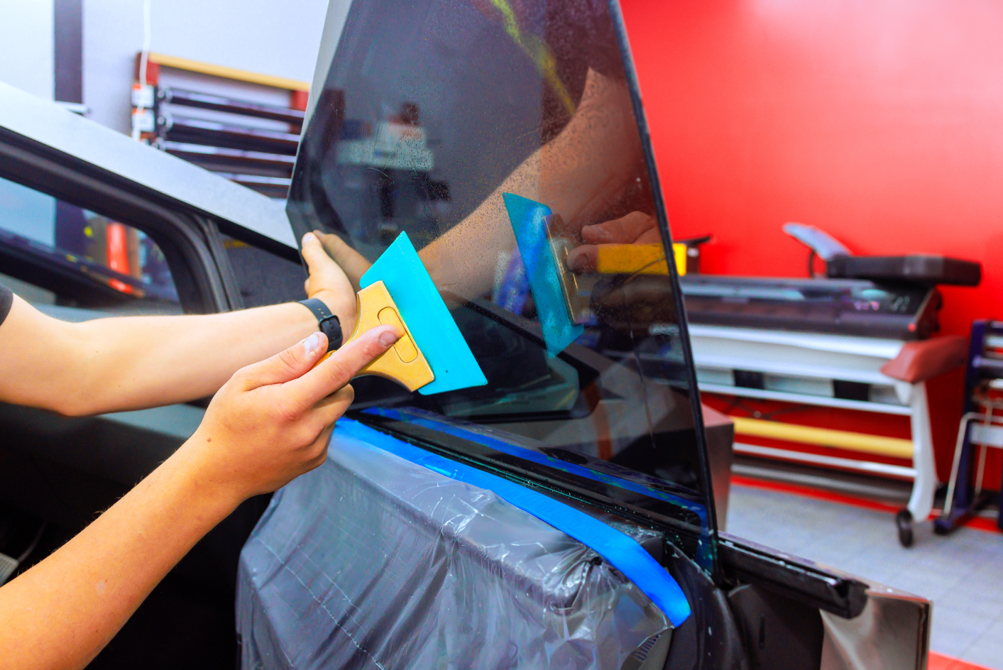 Person applying tinted film to a car window with a squeegee in a workshop setting.