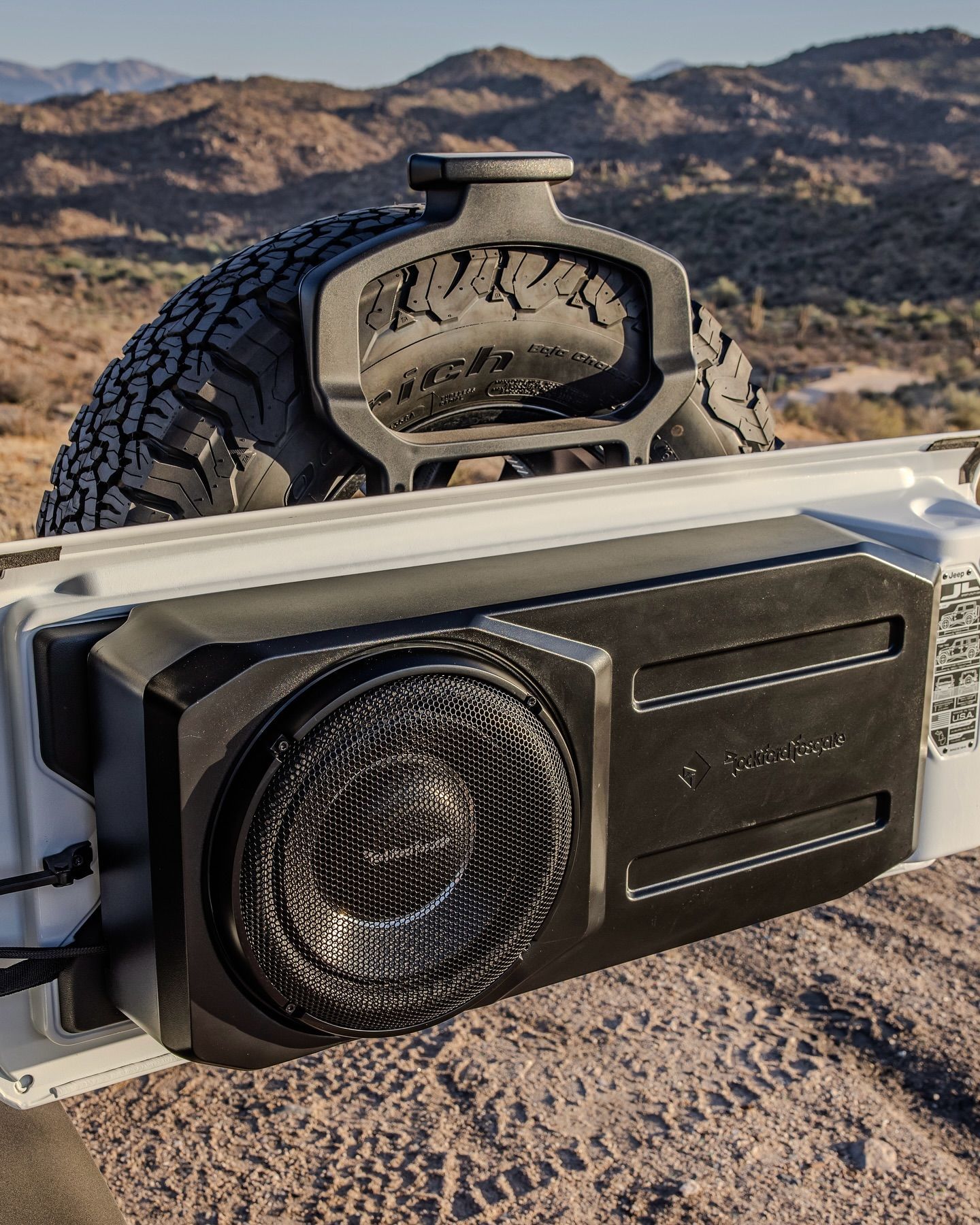Black subwoofer mounted on a white vehicle door, with a spare tire above it and mountains in the background.