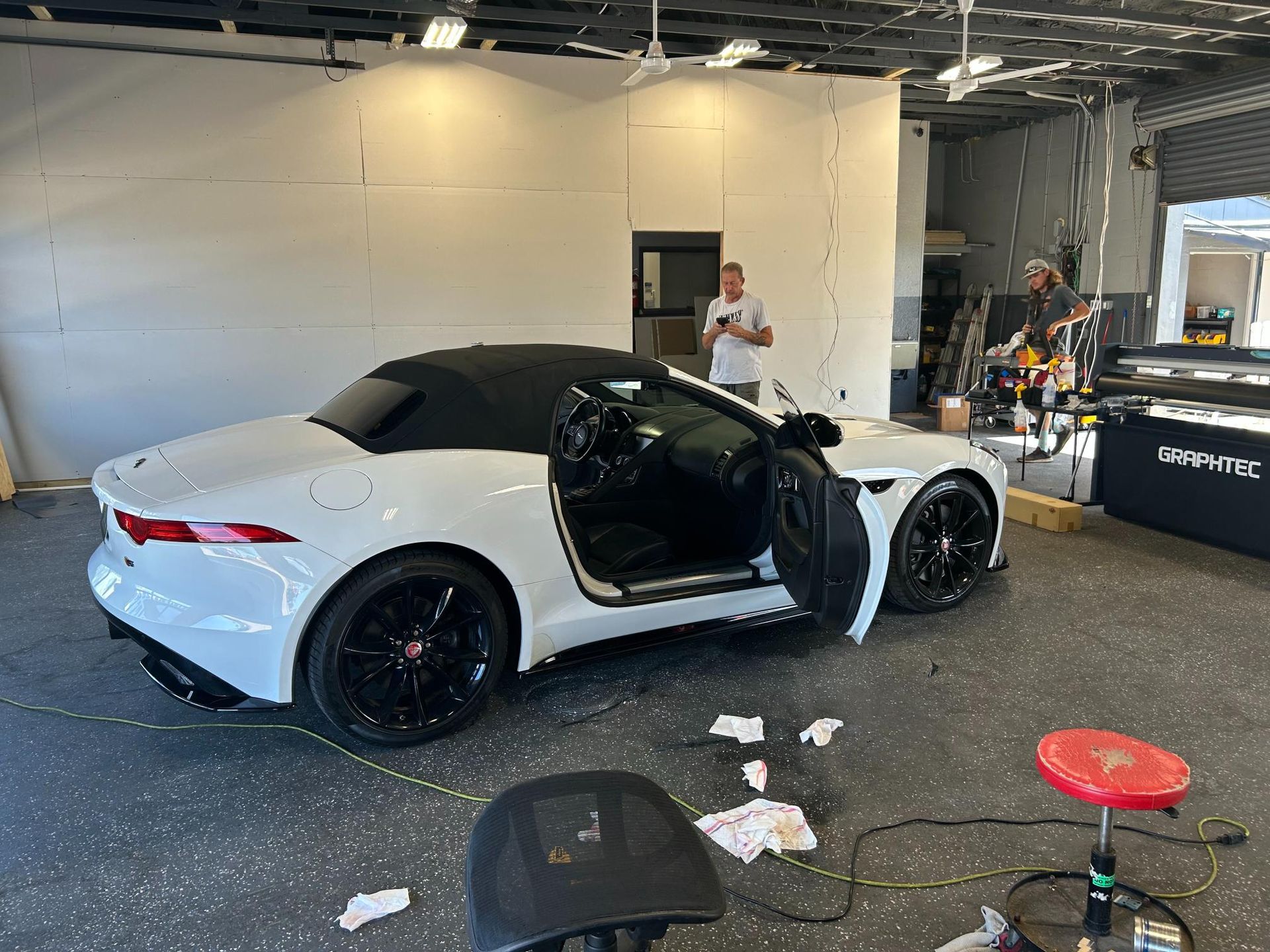 White Jaguar convertible with black wheels, door open in a workshop. Person holding a phone nearby.