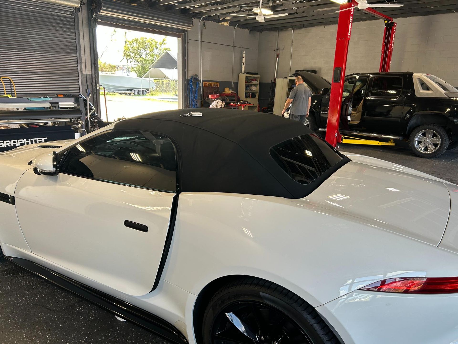 White convertible car in a garage with black top. A person works on another vehicle in the background.