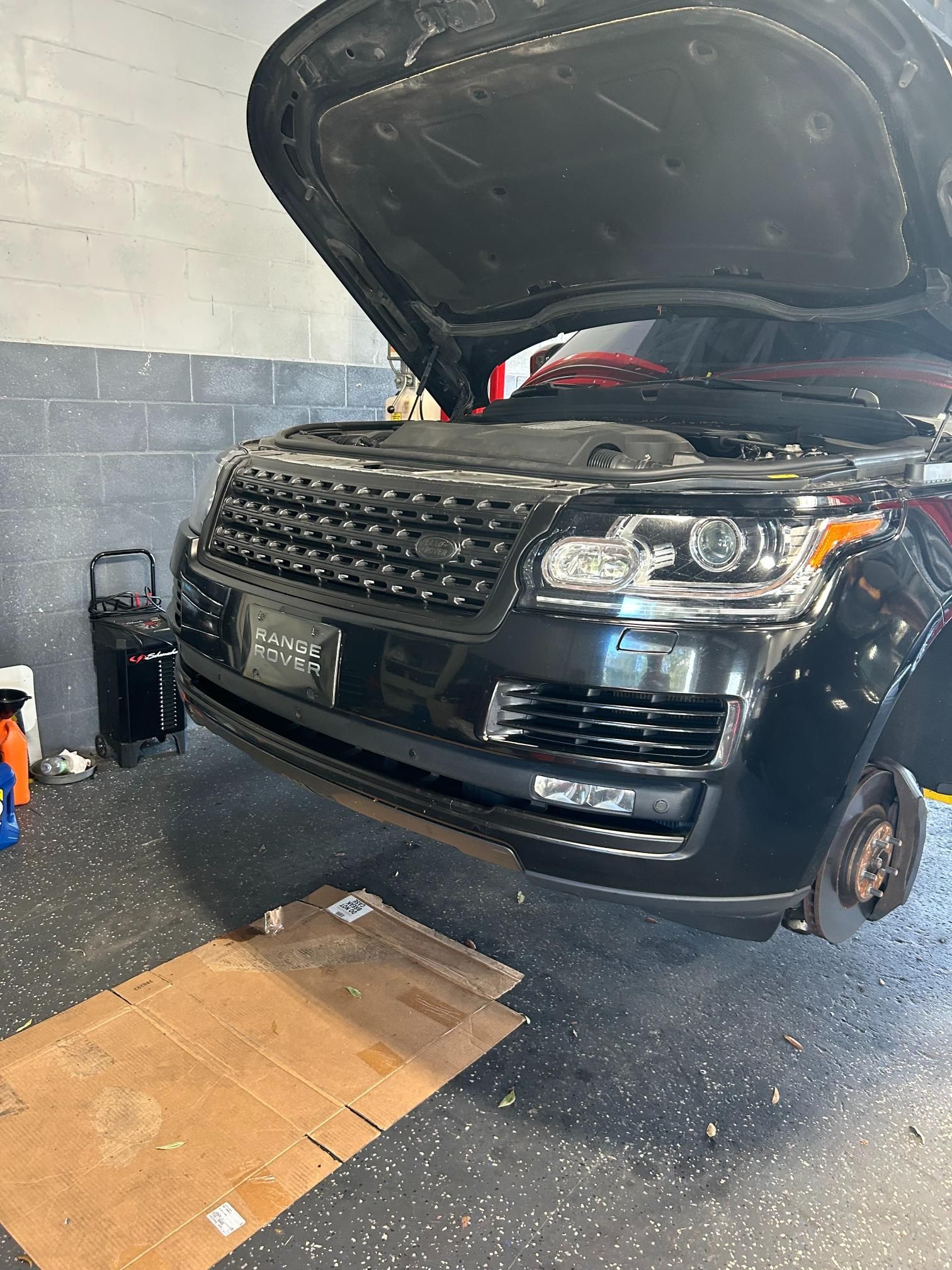 Black Range Rover with the hood open in a repair shop; front wheel removed.