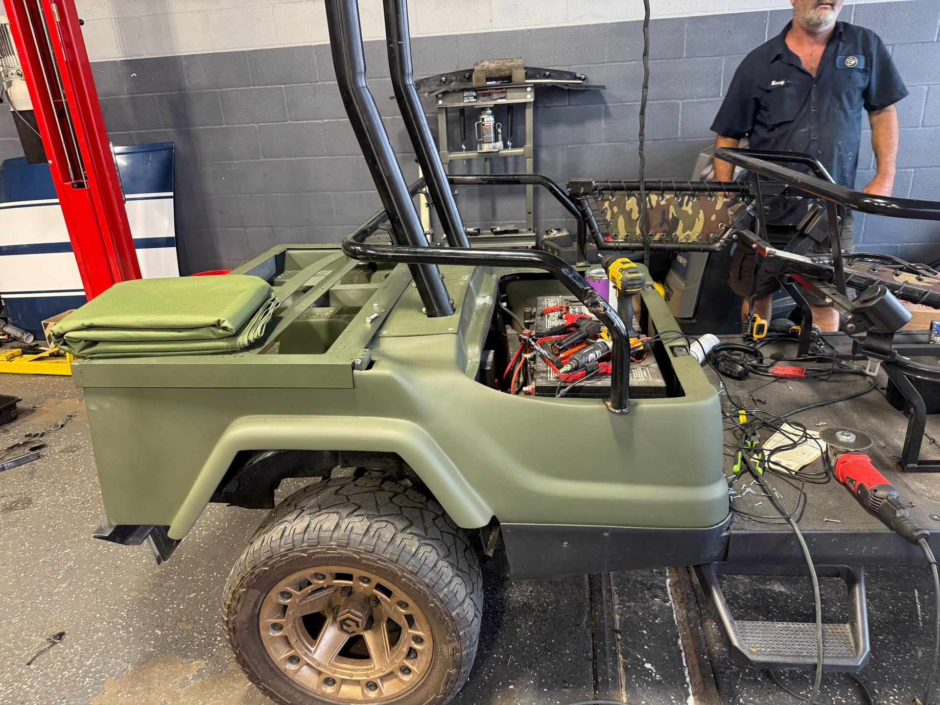 An olive green golf cart chassis being worked on in a garage; a man stands near it.