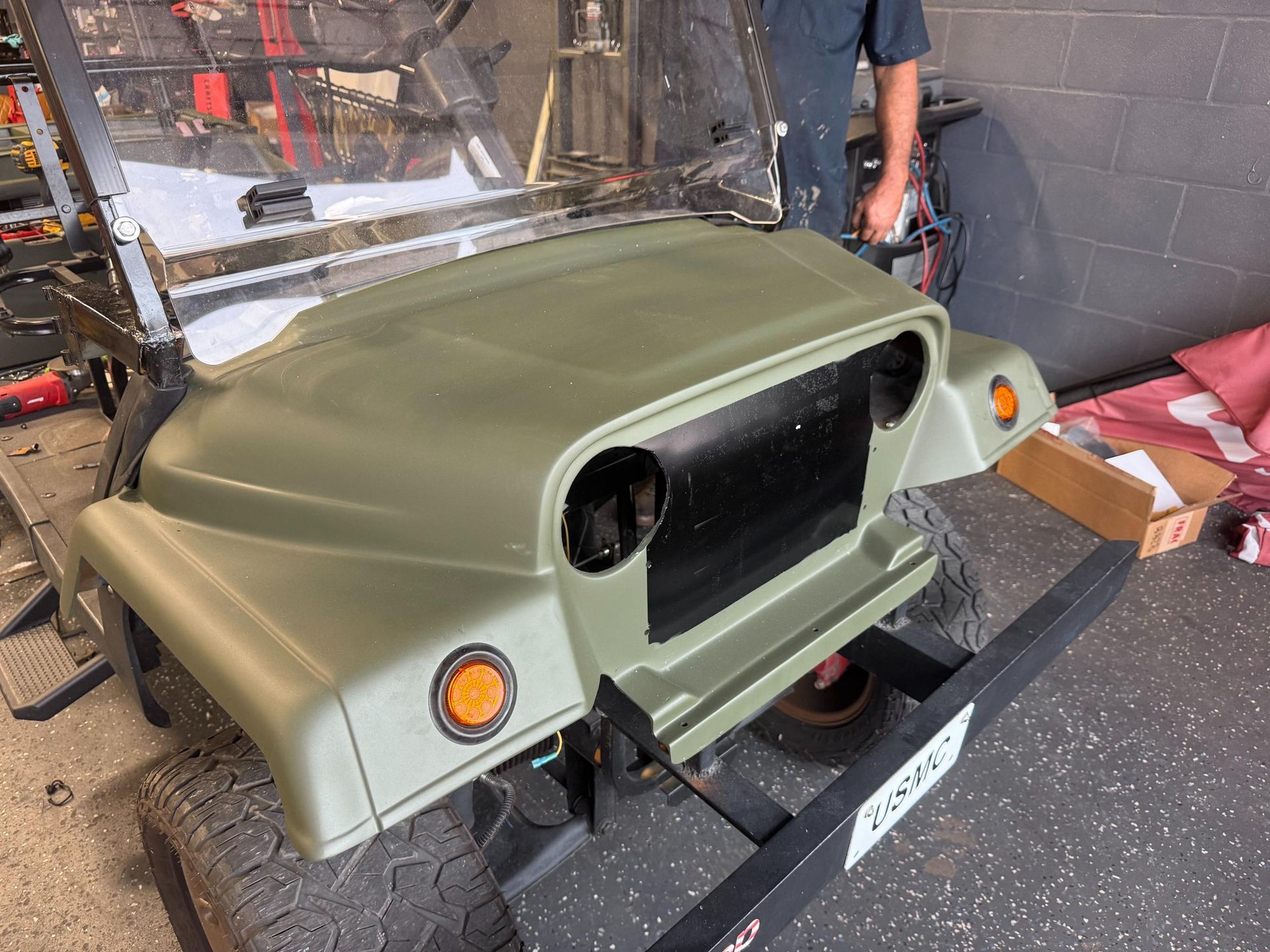 Green golf cart hood with black grill, orange lights, and two people working on it.