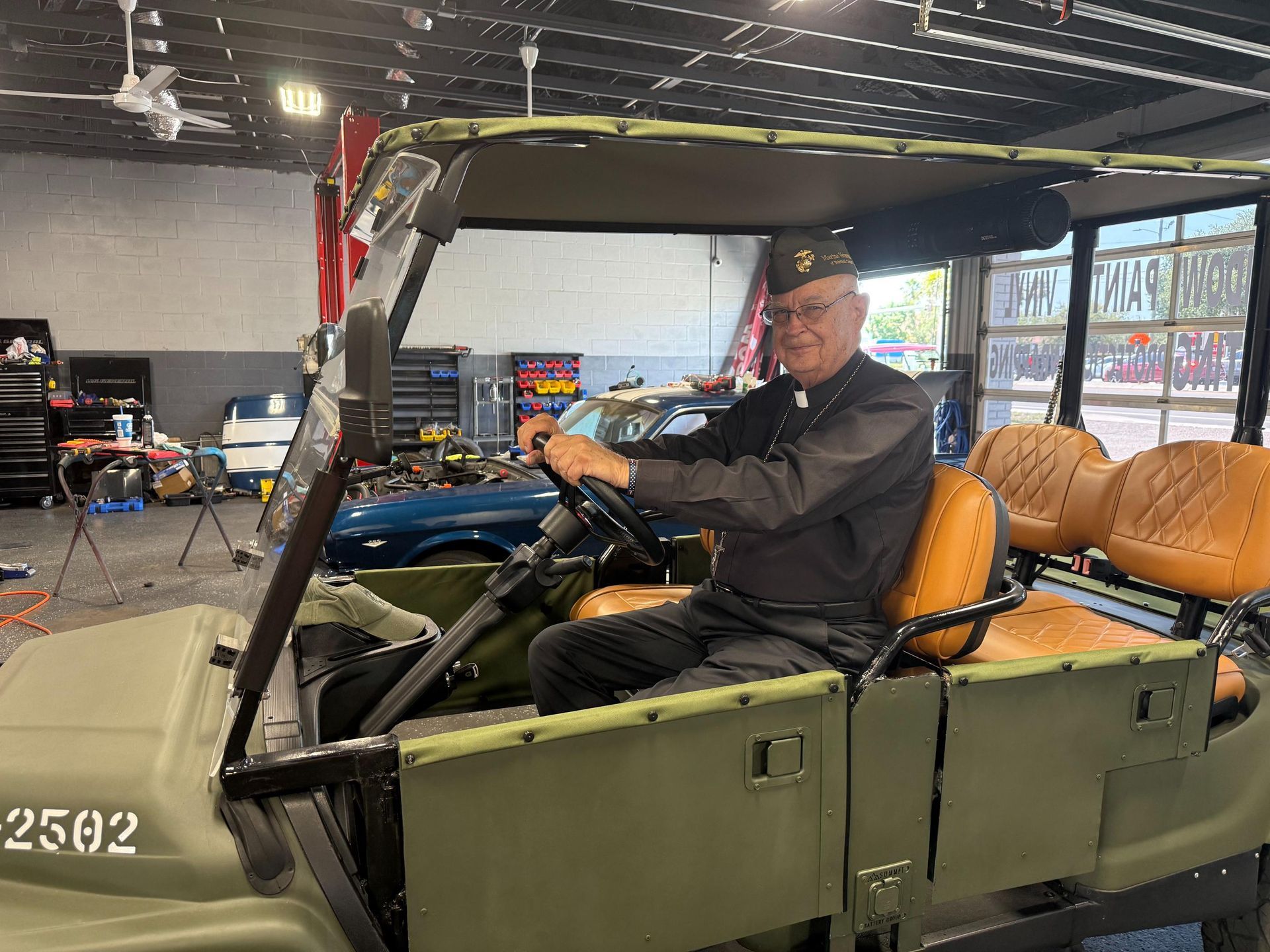 Man in clerical collar and hat smiles, driving a green golf cart in a garage.