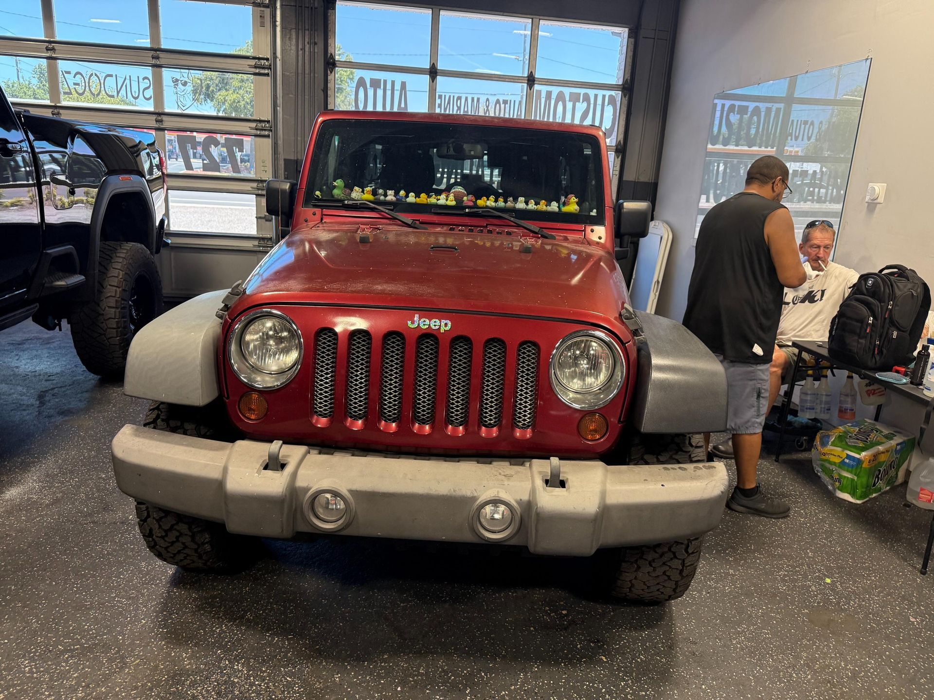 Red Jeep Wrangler with custom grill, parked inside a garage. Two men are present.