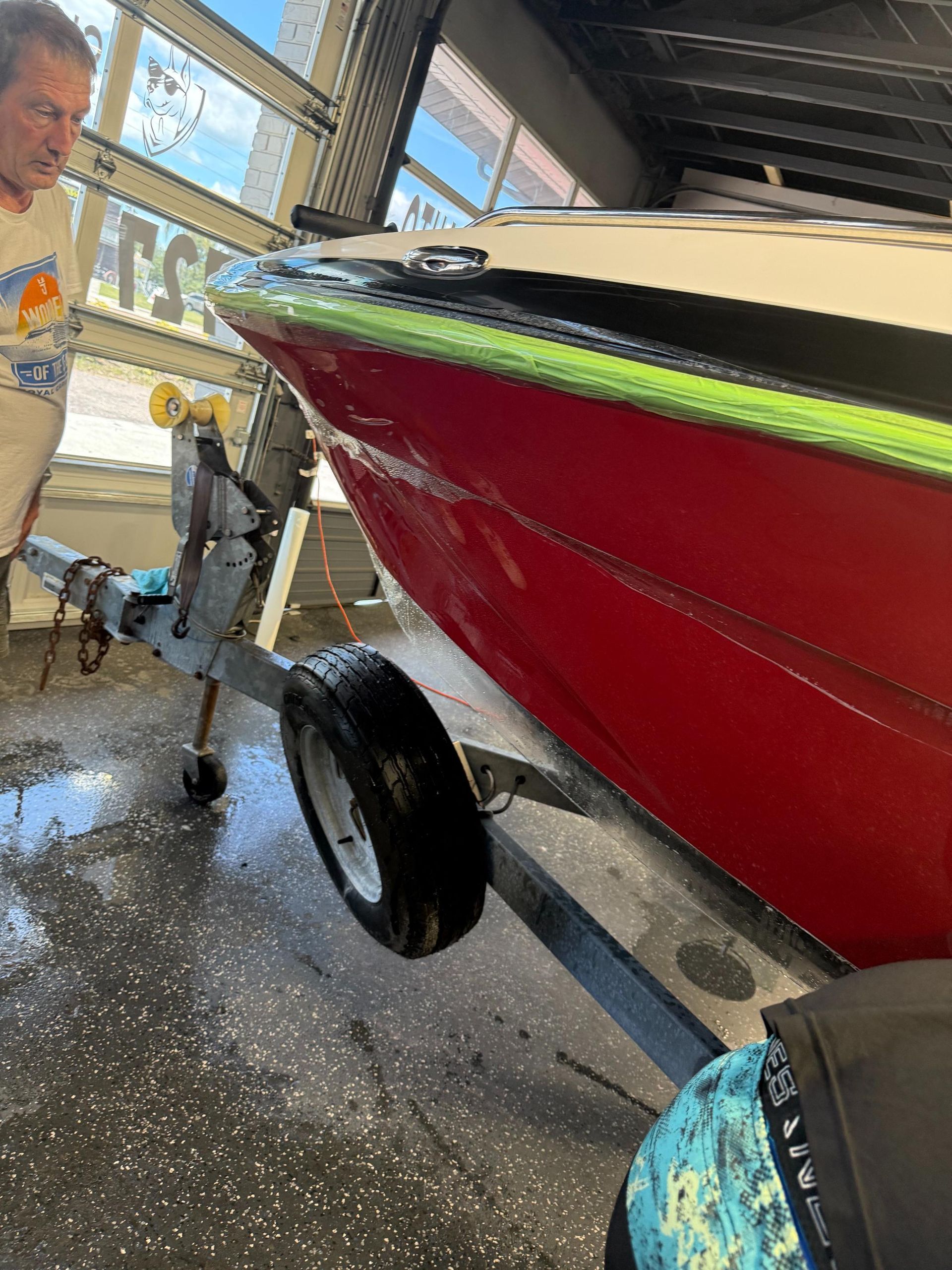 Red boat on trailer; person washes it, green trim, sunny setting.