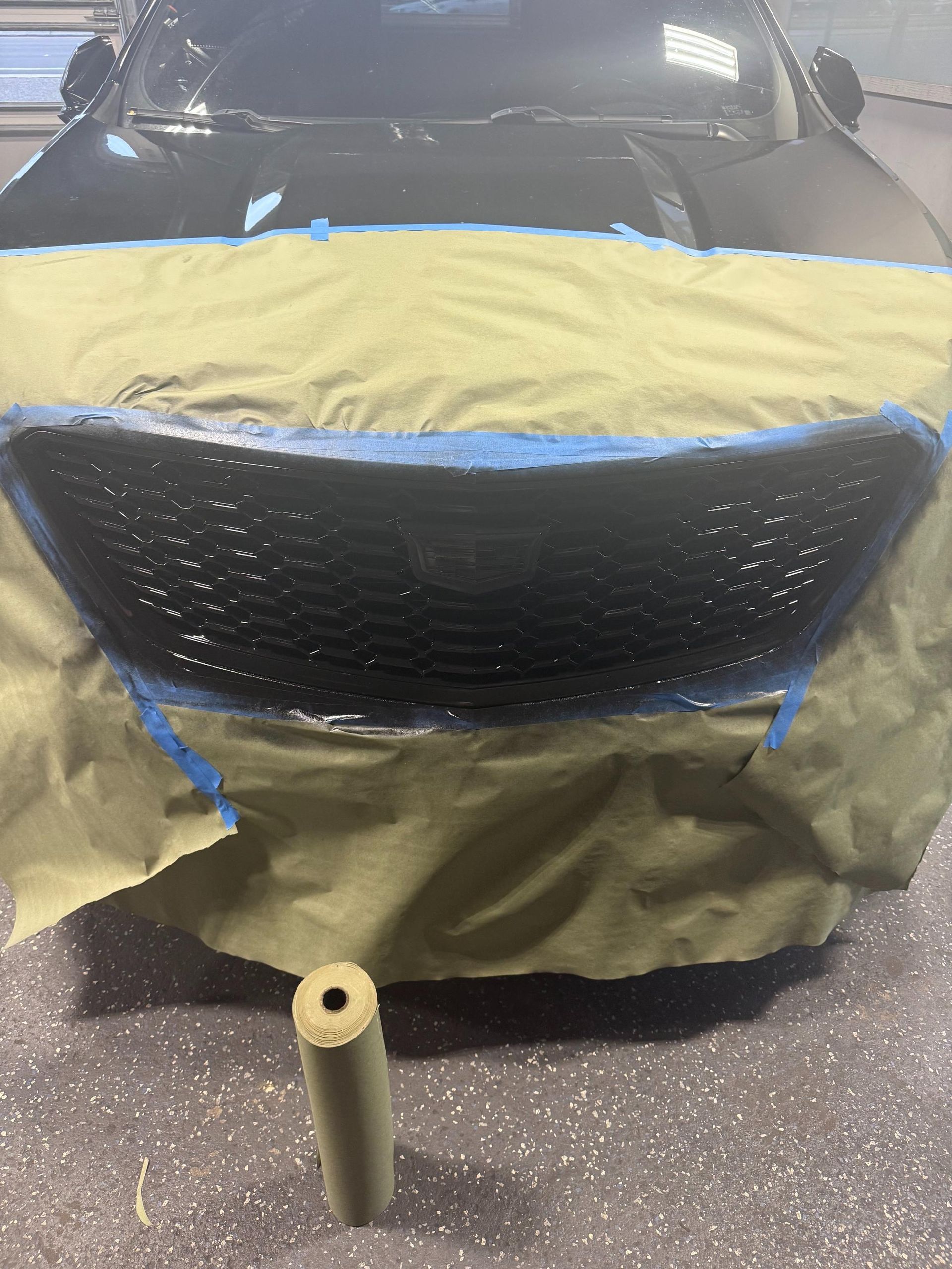 Car grille being painted black, masked with tape and paper, roller in front.