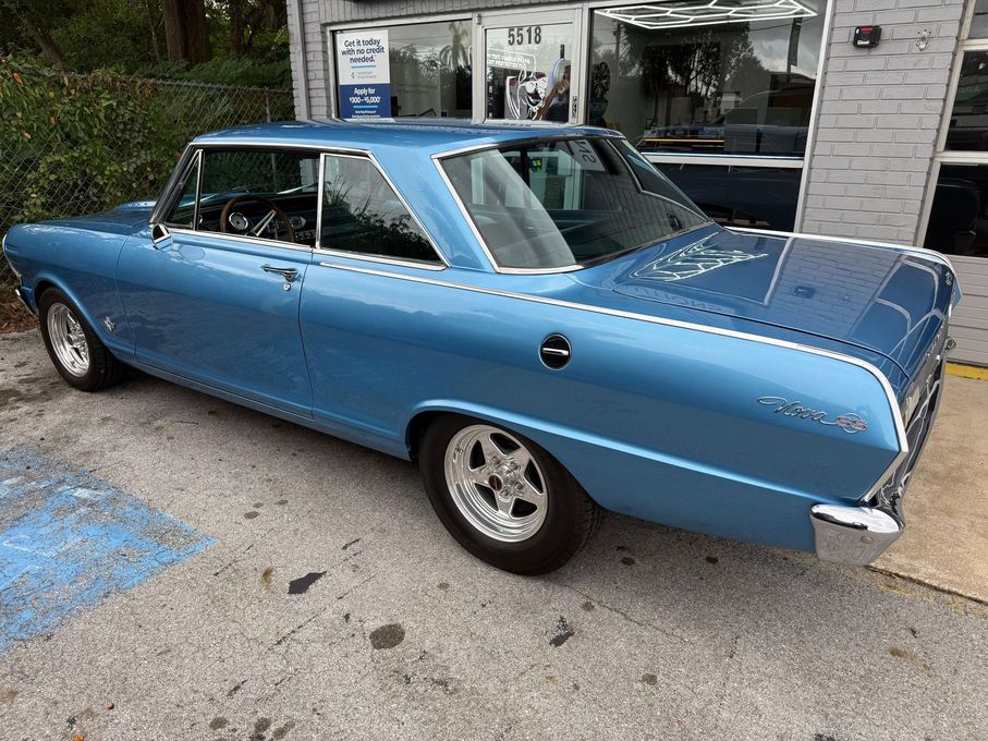 Blue 1965 Chevrolet Nova coupe parked outside a shop. Chrome trim, mag wheels.